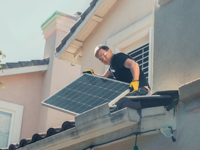 Man Wearing Safety Glasses and Gloves Holding Solar Panels on the Roof